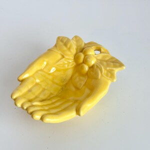 May include: A bright yellow ceramic trinket dish shaped like two cupped hands held together. The wrist area features a decorative relief of three small round berries nestled among several sculpted leaves, creating a textured surface.