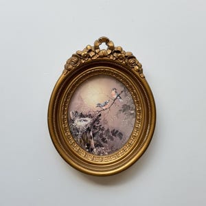 May include: Oval, gold-colored framed artwork featuring a painting of birds perched on a branch near a nest. The frame has a decorative floral design at the top. The artwork is set against a light background.