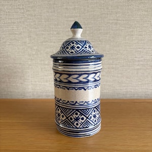May include: A cylindrical ceramic canister with a matching lid, featuring a speckled off-white base decorated with intricate hand-painted blue geometric patterns, including cross-hatching, horizontal bands, and a pointed finial on the lid.