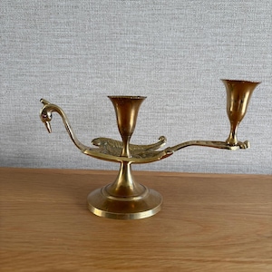 May include: A brass dual-candle holder shaped like a swan with a long neck, a small red stone eye, and two fluted candle cups. The base is circular and supports the central body, which features engraved wing details and an extended arm for the second cup.