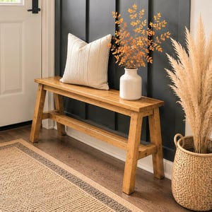 May include: A golden oak wooden bench with a rectangular seat and four angled legs. A cream-colored throw pillow sits on the bench next to a white vase containing orange foliage. The text Golden Oak is visible on the dark wall behind the bench.