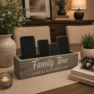 May include: Weathered gray wooden phone organizer with the words "Family Time" and "Leave All Electronics Here" in white script. The organizer holds four smartphones. A candle, books, and decorative items are also on the table.