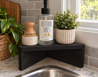 May include: A black wooden corner shelf featuring a clear soap dispenser with a black pump, labeled Autumn Vibes with sunflower graphics, a wooden scrub brush in a speckled ceramic holder, and a small green plant in a ribbed pot.