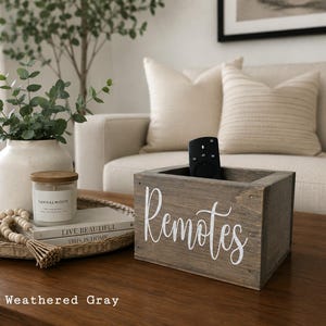 May include: A rectangular weathered gray wooden storage box with the word Remotes written in white cursive script on the front. The box is open at the top and contains a black television remote control. The wood has a visible grain texture.