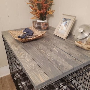 May include: A dog crate with a weathered gray wooden top. The top is made of wooden planks and sits on top of a black wire crate. A woven basket, a framed photo, and decorative flowers are on the top.