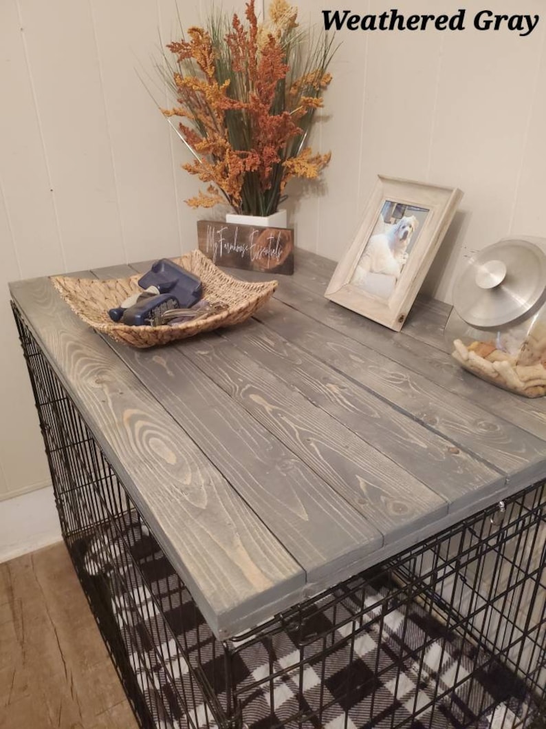 May include: A dog crate with a weathered gray wooden top. The top is made of wooden planks and sits on top of a black wire crate. A woven basket, a framed photo, and decorative flowers are on the top.