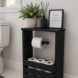 May include: A black wooden bathroom storage unit with a shelf holding a potted plant, a black diffuser, and a framed sign that reads "WASH YOUR HANDS." The unit also has a toilet paper holder and storage for extra rolls.