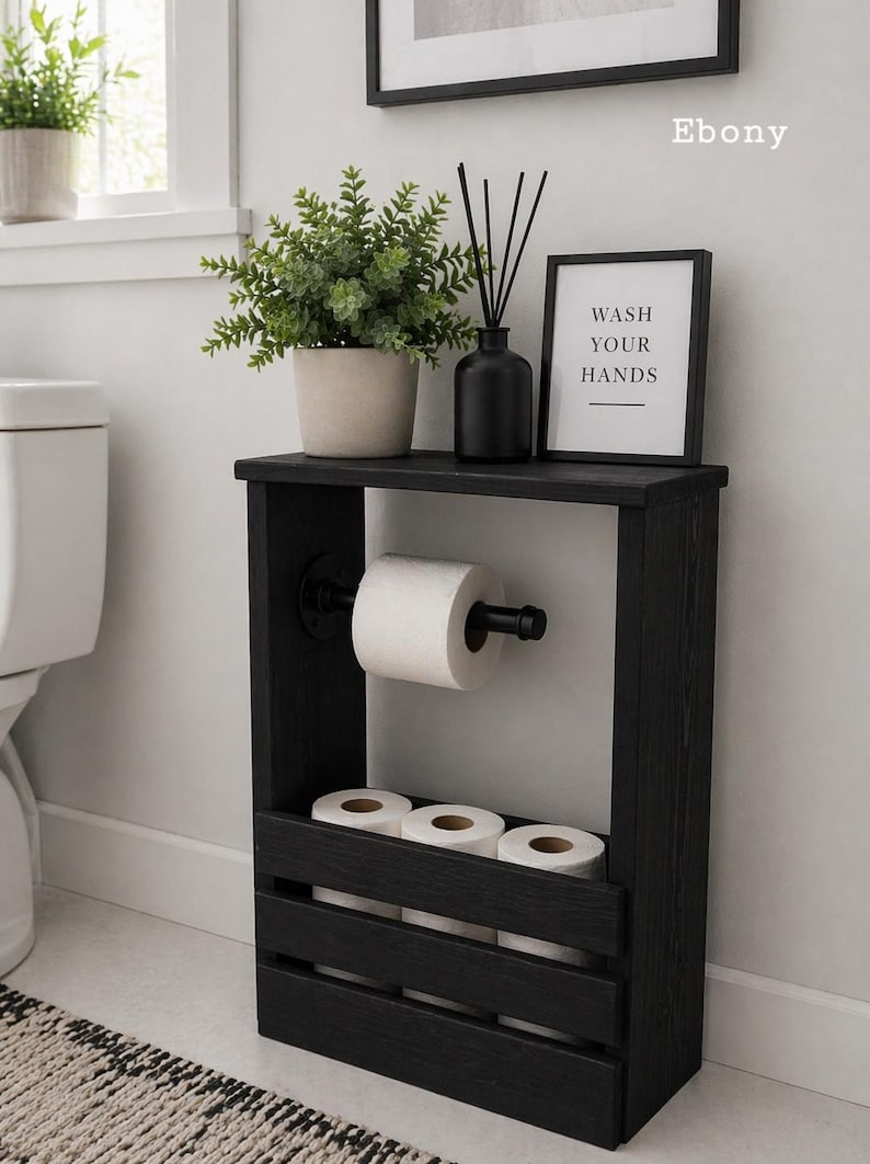 May include: A black wooden bathroom storage unit with a shelf holding a potted plant, a black diffuser, and a framed sign that reads "WASH YOUR HANDS." The unit also has a toilet paper holder and storage for extra rolls.