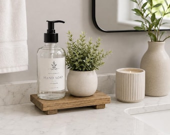 May include: A clear glass hand soap dispenser with a black pump, a white label reading "Premium Quality Hand Soap Lavender 16 oz | 475 ml", and a small green plant in a beige pot, all resting on a rectangular wooden riser labeled "4x8 Early American".