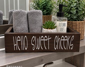 May include: A dark brown wooden rectangular storage box with the white text Hello Sweet Cheeks printed on the front. Inside the box are two rolled gray towels, a small green succulent in a white pot, and a clear soap dispenser with the text Autumn Vibes.