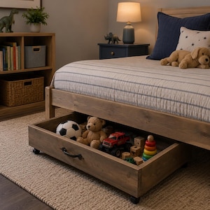May include: A wooden bed frame with a pull-out storage drawer underneath, featuring a black metal handle and wheels. The drawer contains a soccer ball, a teddy bear, a red toy truck, wooden blocks, and a colorful stacking ring toy.