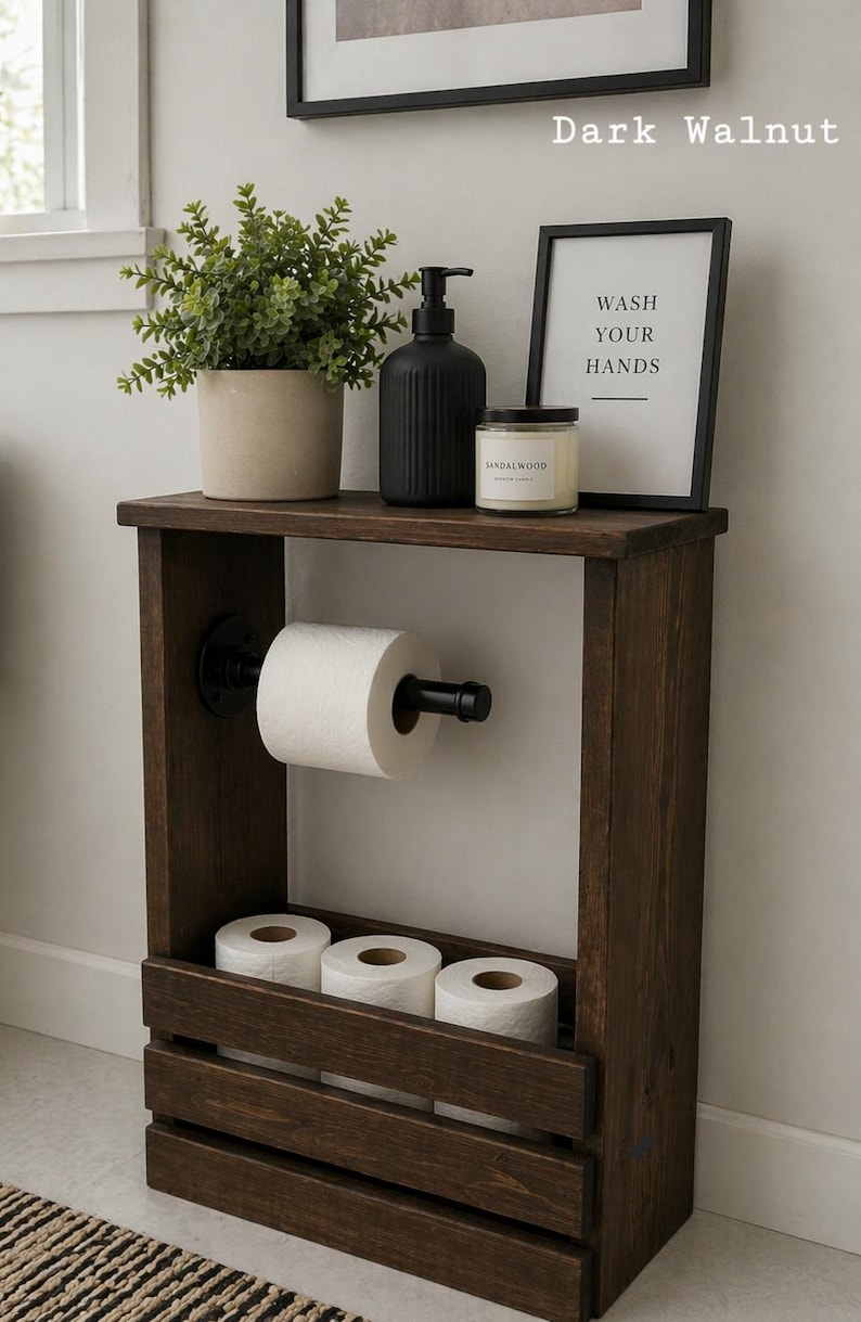 May include: Dark walnut wooden bathroom storage unit with a shelf holding a potted plant, black soap dispenser, and a candle. The unit has a toilet paper holder and a lower shelf with three rolls of toilet paper. A framed sign reads "WASH YOUR HANDS".