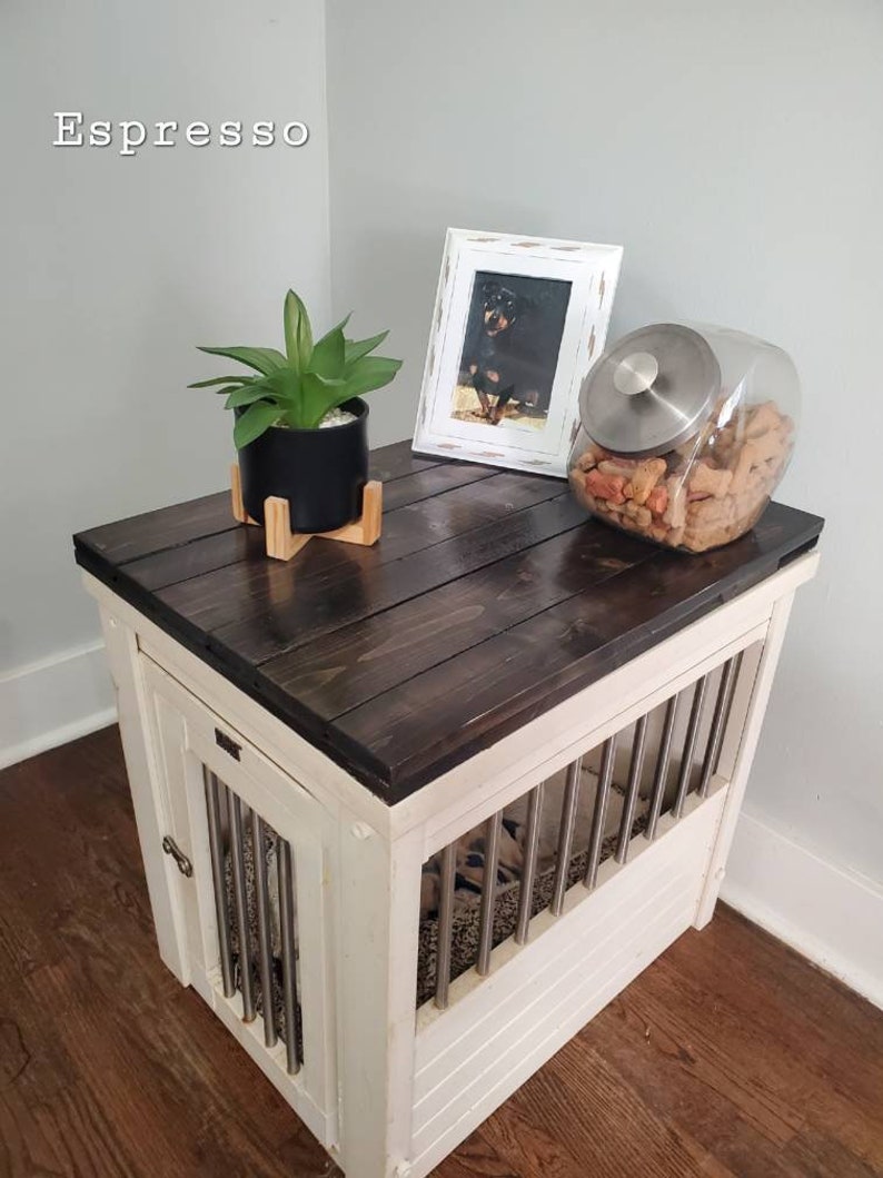 May include: Espresso dog crate with a dark stained top and white base. Features metal bars on the front and a door with a latch. The top holds a framed photo, a plant, and a glass jar of dog treats.
