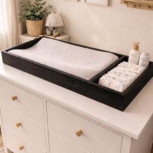 May include: A black wooden changing table organizer with a white changing pad, diapers, and baby products. The organizer sits on a white dresser with gold knobs. The word "Ebony" is visible on the bottom right.