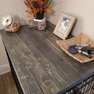 May include: Carbon Gray dog crate with a wooden top. The top is stained a dark gray color and has a natural wood grain pattern. A glass jar with dog treats, a framed photo of a dog, and a woven basket with grooming tools are on top.