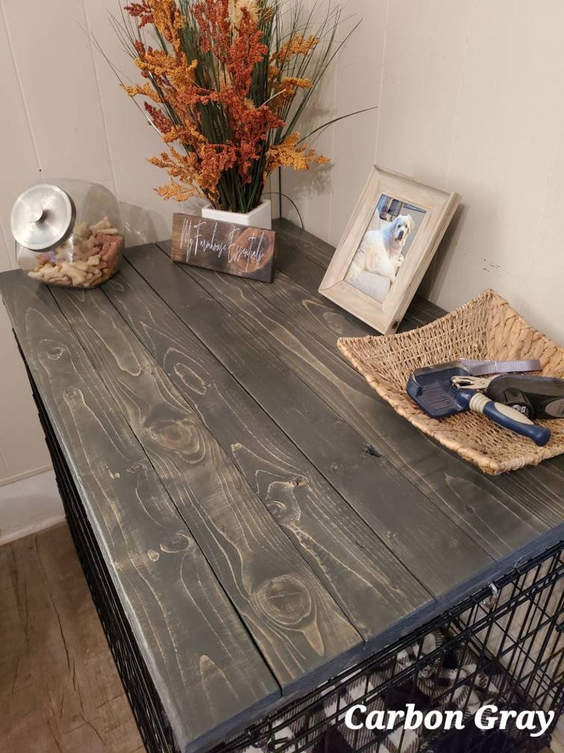 May include: Carbon Gray dog crate with a wooden top. The top is stained a dark gray color and has a natural wood grain pattern. A glass jar with dog treats, a framed photo of a dog, and a woven basket with grooming tools are on top.