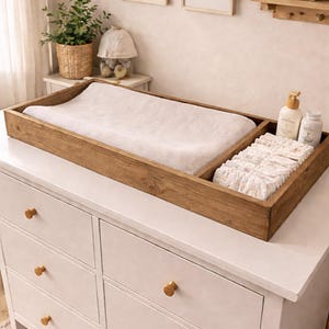May include: A wooden changing table topper with a white changing pad and a section for diapers and baby products. The topper sits on a white dresser with gold knobs. The text "Special Walnut" is at the bottom.