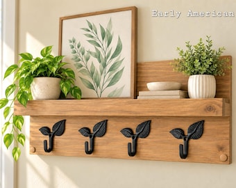 May include: A wooden wall-mounted shelf with four black metal hooks shaped like leaves. The shelf features a light brown wood grain finish and the text Early American. It holds a potted trailing plant, a framed botanical print, and a small plant.