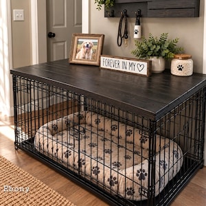 Può includere: Una gabbia per cani in metallo nero con un piano superiore in legno scuro. All'interno si trova una cuccia beige con un motivo a impronte nere. Sul piano in legno poggiano una cornice, un cartello con scritto "FOREVER IN MY ♡", una pianta e un barattolo.