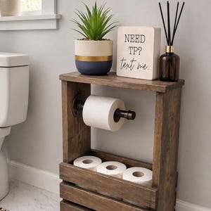 May include: A rustic wooden toilet paper holder with a shelf, holding a roll of toilet paper and three additional rolls. A small potted plant, a sign that says "NEED TP? text me," and a diffuser bottle are on top.
