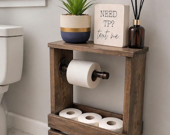 May include: A dark brown wooden toilet paper holder stand with a metal pipe arm for one roll and a lower crate section for three additional rolls. On top sits a potted green plant, a reed diffuser, and a sign that reads NEED TP? text me.