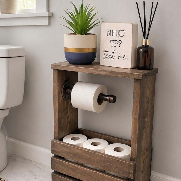 May include: A dark brown wooden toilet paper holder stand with a metal pipe arm for one roll and a lower crate section for three additional rolls. On top sits a potted green plant, a reed diffuser, and a sign that reads NEED TP? text me.