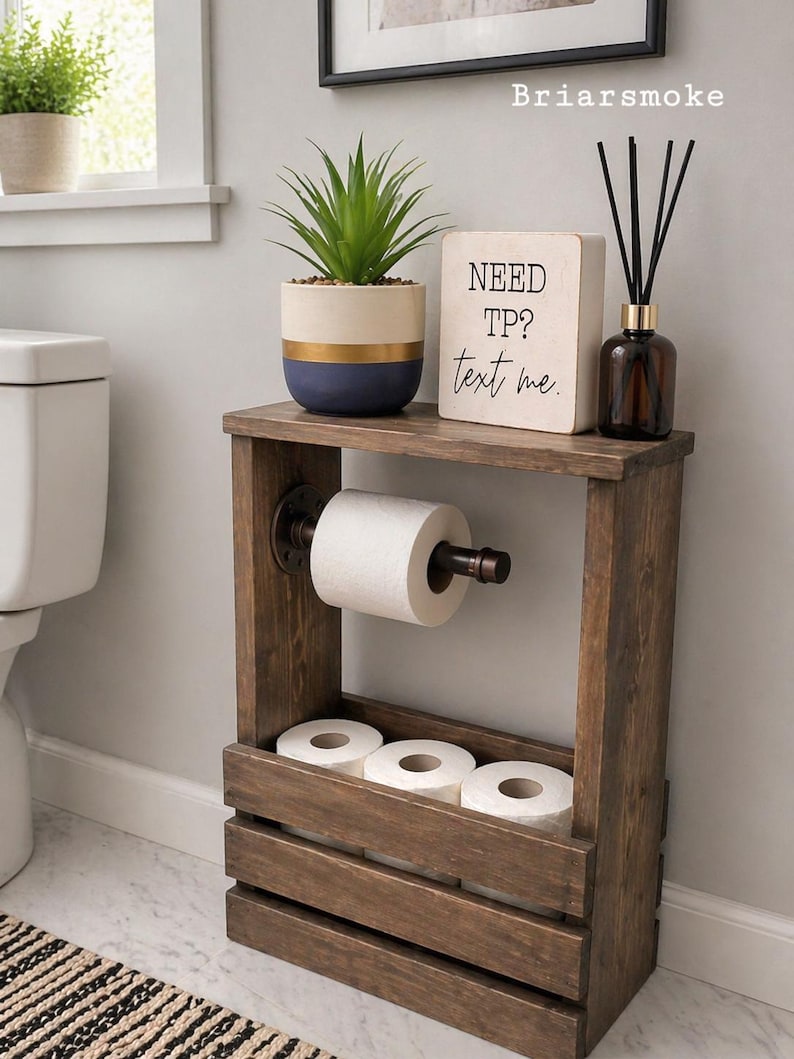 May include: A rustic wooden toilet paper holder with a shelf, holding a roll of toilet paper and three additional rolls. A small potted plant, a sign that says "NEED TP? text me," and a diffuser bottle are on top.