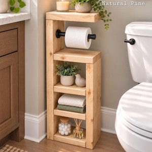 Può includere: Un mobiletto da bagno in legno di pino naturale con portarotolo in metallo nero, tre ripiani aperti e la scritta Natural Pine. I ripiani ospitano una piccola pianta, una statuetta di uccellino, asciugamani piegati, un barattolo di cotone e un diffusore di fragranze.