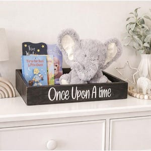 May include: A decorative nursery scene featuring a black wooden tray with the text "Once Upon A Time", holding books and a plush elephant. A white lamp, rainbow stacker, and framed art complete the display.