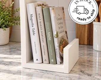 May include: A white wooden cookbook stand holding four books titled Magnolia Table, Half Baked Harvest, Skinnytaste, and The Complete Cooking for Two Cookbook. The stand has a simple L-shaped design with a flat base and two vertical side supports.