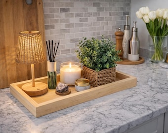 May include: A rectangular light-colored wooden tray holding a small woven lamp, a reed diffuser with black sticks, a white candle in a glass jar, a small purple stone, a round container, and a green plant in a woven square basket.