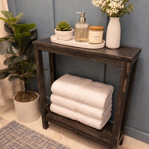 May include: A dark stained wooden bathroom shelf with a stack of white towels on the bottom shelf. The top shelf holds a small succulent, soap dispenser, candle, and a vase of white flowers. A potted plant is to the left.