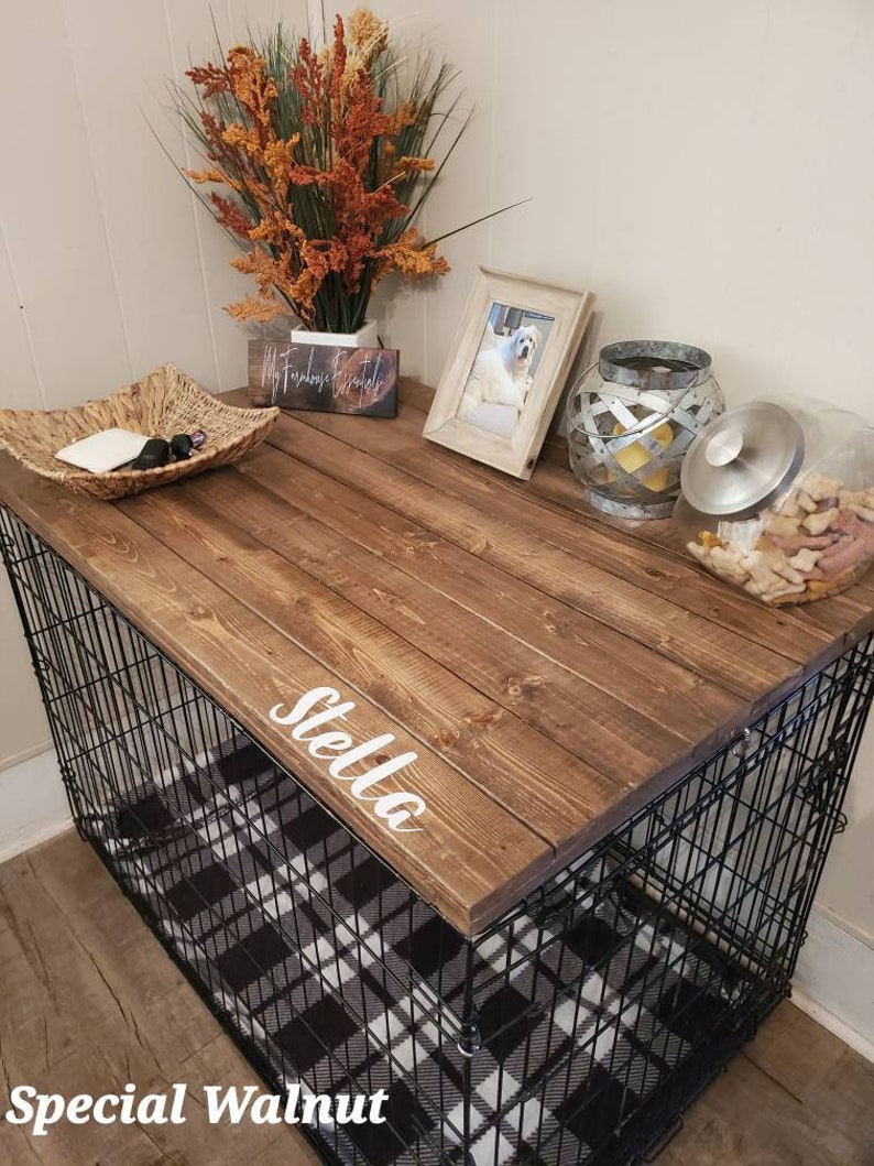 May include: A dog crate with a wooden top and a black wire frame. The top is stained a dark brown and has the name "Stella" in white script. The crate sits on a black and white plaid bed. Decorative items include a photo frame and a glass jar.