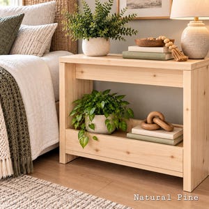 May include: A rectangular natural pine console table featuring a flat top surface and a lower shelf. The table has a light wood grain finish and is styled with a potted green plant, stacked books, a wooden bowl, and a decorative wooden chain link.