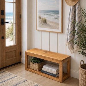 May include: A rectangular golden oak wooden bench with a lower shelf holding a woven basket and three stacked books titled Coastal Home, Live by the Water, and The Kinfolk Home, positioned in an entryway with a beach scene wall art piece.