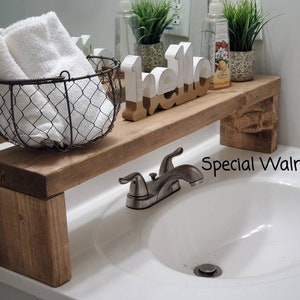 Può includere: Una mensola in legno sopra il lavandino con finitura Special Walnut, dotata di un cestino in filo metallico con asciugamani bianchi, una scritta "hello" bianca e color legno e due piccole piante in vaso sopra il rubinetto.