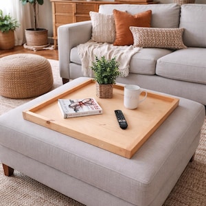 May include: A light gray ottoman with a wooden tray holding a book, remote, and mug. The tray is surrounded by a light gray sofa with decorative pillows. The room is decorated with plants and a woven rug.
