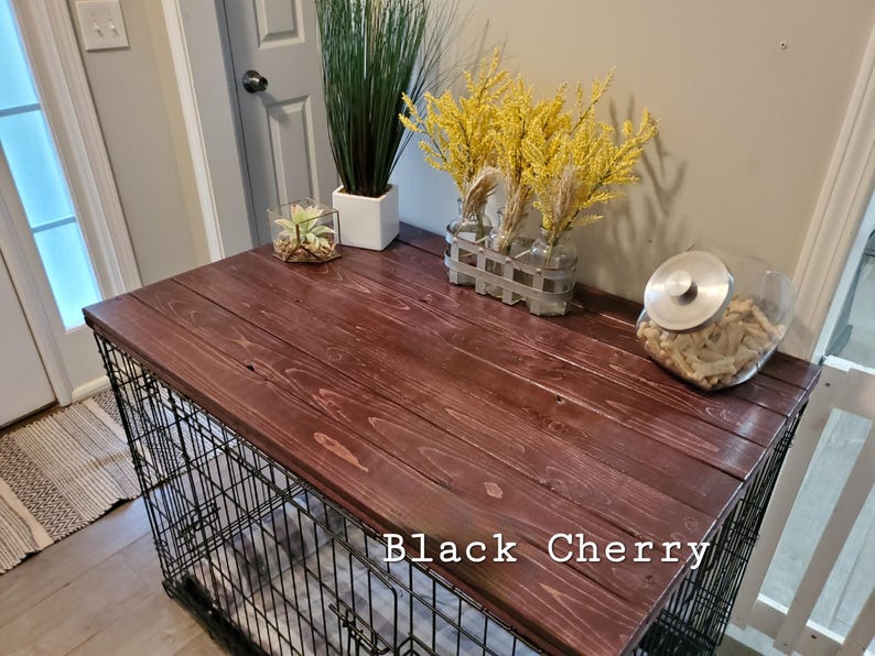 Pu&ograve; includere: Una copertura in legno marrone scuro per gabbia per cani con finitura liscia e venature del legno visibili, con il testo Black Cherry stampato sulla superficie. La parte superiore &egrave; piatta e rettangolare, usata come piano per piante.
