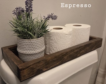 May include: A rectangular dark brown wooden tray holding a gray textured pot with a faux lavender plant and two white toilet paper rolls. The word Espresso is printed in white text above the tray, which sits on a white toilet tank.