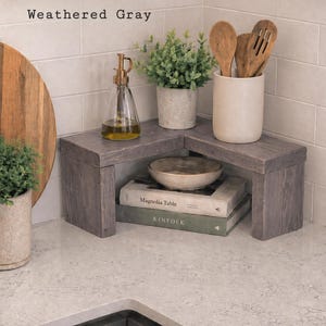 Può includere: Una mensola ad angolo rifinita in legno grigio invecchiato. La mensola sostiene un dosatore per olio in vetro, una piccola pianta in vaso e un portautensili in ceramica con cucchiai di legno. Sotto la mensola ci sono due libri, Magnolia Table e KINFOLK.