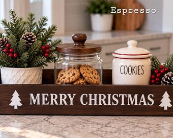 May include: A dark brown wooden rectangular tray with the white text MERRY CHRISTMAS and two white tree icons. It holds a small potted evergreen plant with red berries and pinecones, a glass jar with a wooden lid containing cookies, and a white ceramic cookie jar.