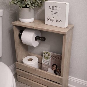 May include: Wooden bathroom storage unit with a toilet paper holder, a magazine rack, and a decorative plant in a gray and white pot. A small white sign reads "NEED TP? text me."