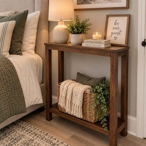 May include: A dark brown wooden console table with four legs and a lower shelf. The top holds a white textured lamp, a potted green plant, a candle, and two stacked books labeled A GOOD NIGHT and SWEET DREAMS. A framed sign reads be our guest.