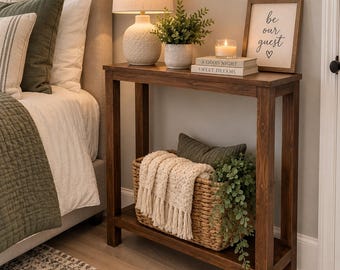 May include: A dark brown wooden console table with four legs and a lower shelf. The top holds a white textured lamp, a potted green plant, a candle, and two stacked books labeled A GOOD NIGHT and SWEET DREAMS. A framed sign reads be our guest.