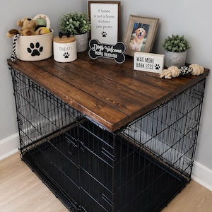 May include: A dog crate with a dark walnut stained wooden top and a black wire frame. The top is decorated with dog-themed items, including a toy, a treat jar, and framed signs that read "A house is not a home without paw prints" and "Wag more, bark less."