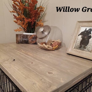 May include: A dog crate with a Willow Gray wooden top. The top has a framed photo of a dog, a glass jar filled with dog treats, and a decorative floral arrangement. The crate has a black metal cage.