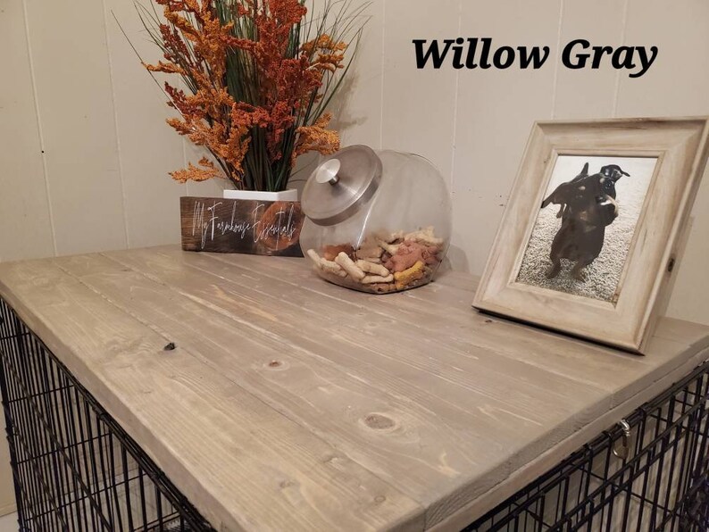 May include: A dog crate with a Willow Gray wooden top. The top has a framed photo of a dog, a glass jar filled with dog treats, and a decorative floral arrangement. The crate has a black metal cage.