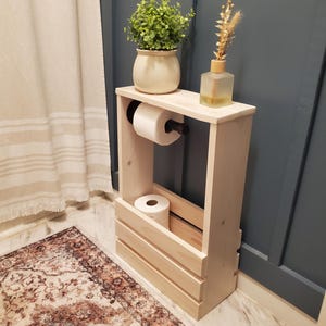 May include: White-washed wooden bathroom storage unit with a shelf holding a potted plant, a decorative bottle, and a roll of toilet paper. The unit also has a lower compartment for additional toilet paper rolls. The text "White-Washed" is visible at the top.