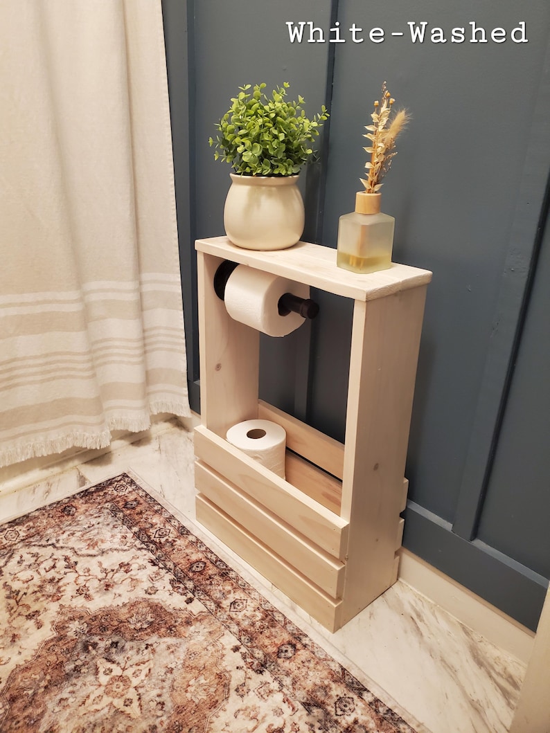 May include: White-washed wooden bathroom storage unit with a shelf holding a potted plant, a decorative bottle, and a roll of toilet paper. The unit also has a lower compartment for additional toilet paper rolls. The text "White-Washed" is visible at the top.
