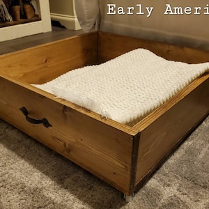 May include: A wooden storage box with a white blanket inside. The box has a black metal handle and is sitting on a light brown rug. The text "Early American" is visible in the background.